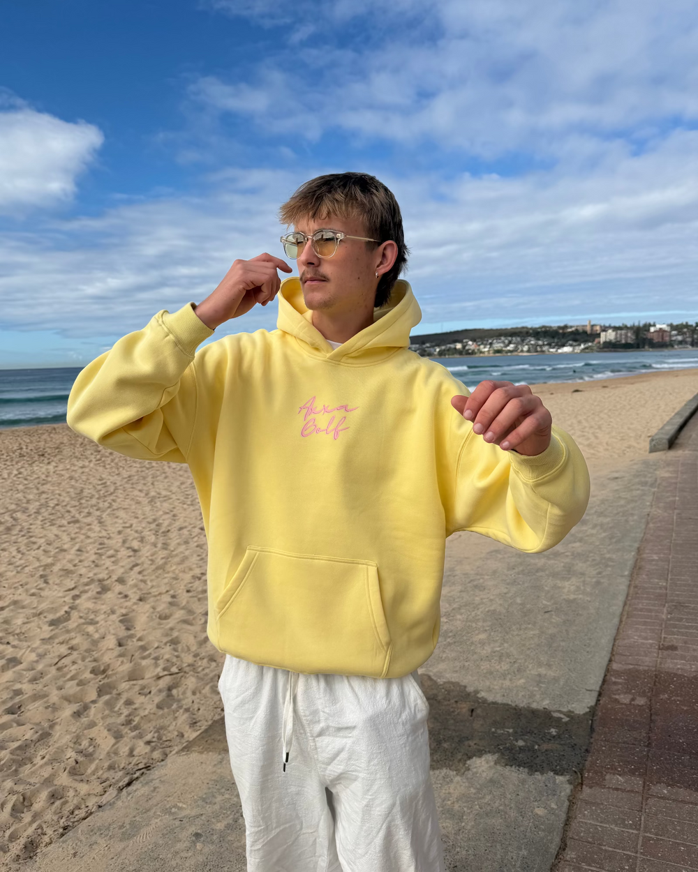 A man wearing a pastel yellow hoodie with a pink graphic on the chest and white sweatpants, standing on a beach boardwalk with sand, ocean, and blue sky in the background.